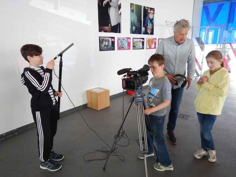 Three young people and one adult using a large professional camera on a stand in modern exhibition room.