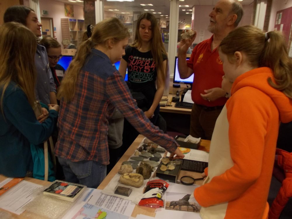 A group of young people gathered around a table covered with pieces of pottery, fossils and information sheets. An adult holds up a fossil to present to the young people.