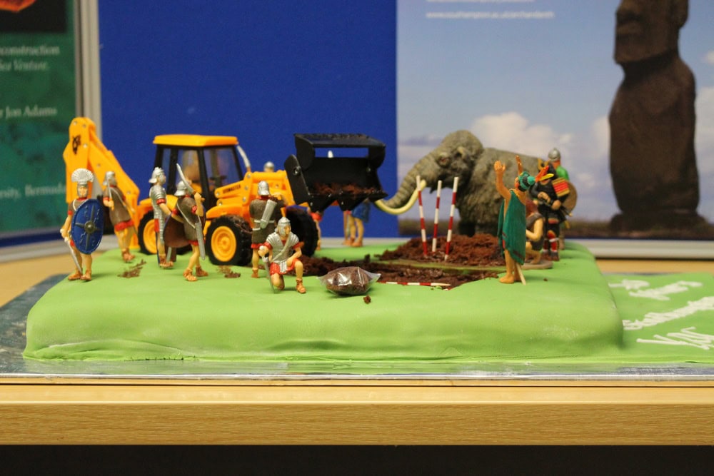 A rectangular cake covered in green icing decorated with plastic models such as a digger, Roman soldiers and a mammoth.