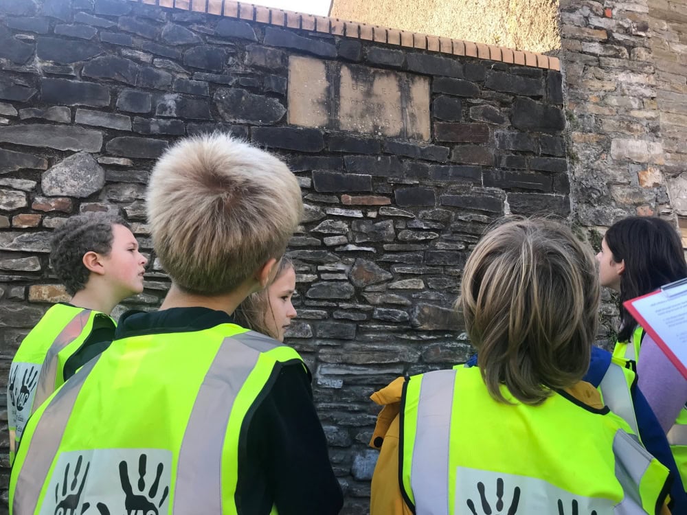 A group of children and adults wearing high-vis jackets with the YAC handprint logo look up at a historic stone wall.