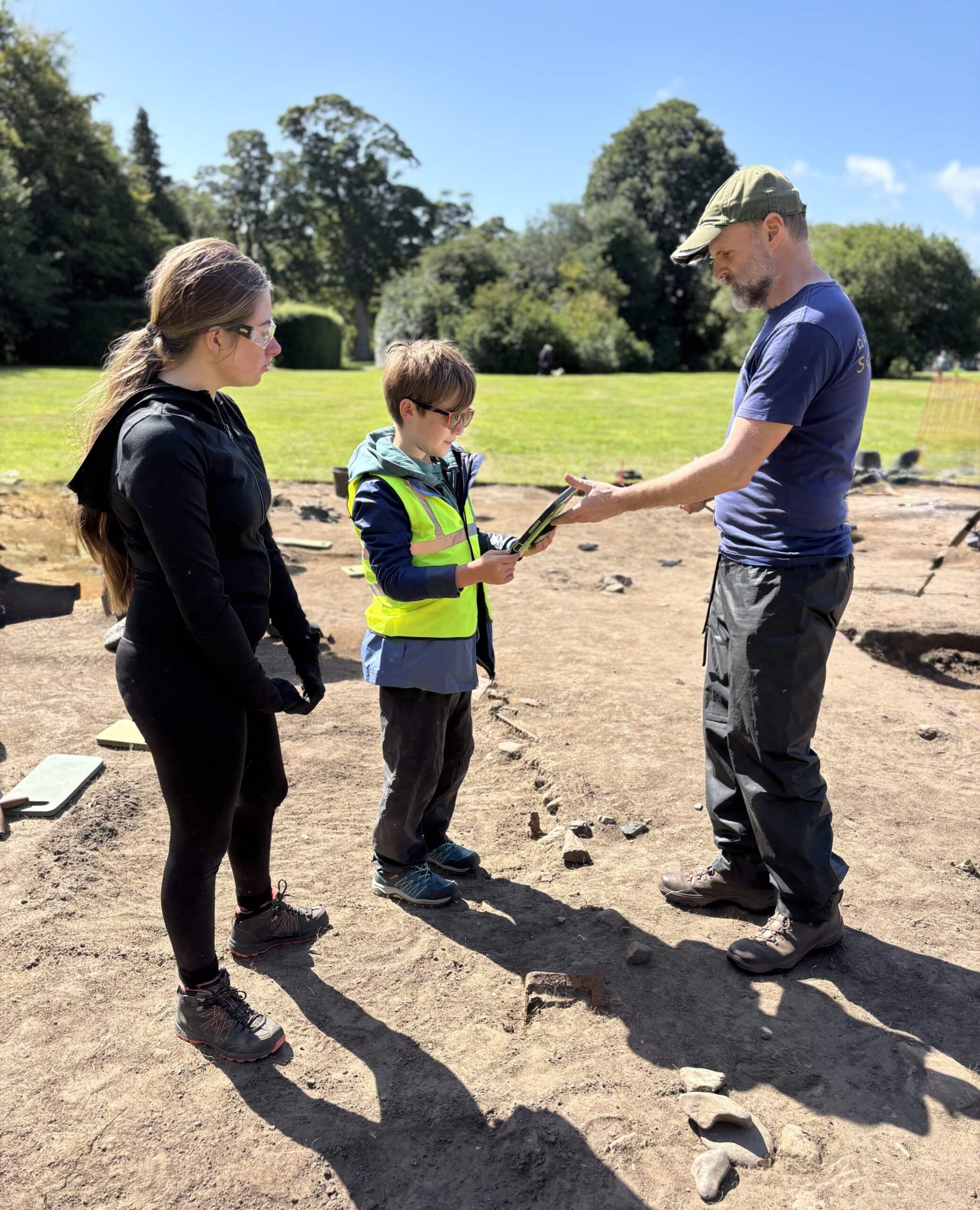 Two young people stand in an archaeological excavation looking at a tablet being held by an adult.