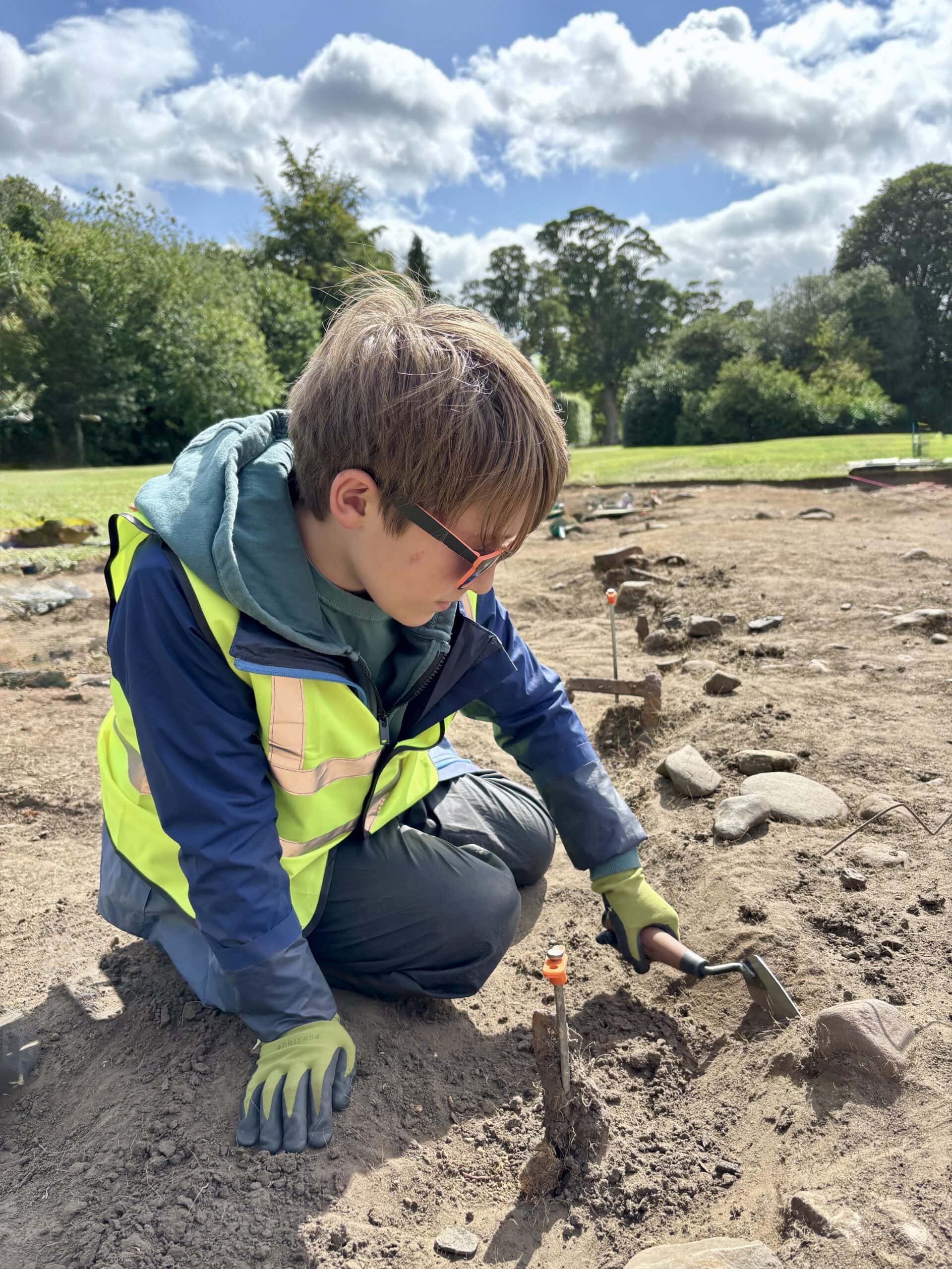 A young person wearing a high-vis vest and gloves kneels in an archaeological trench and scrapes the soil with a trowel.
