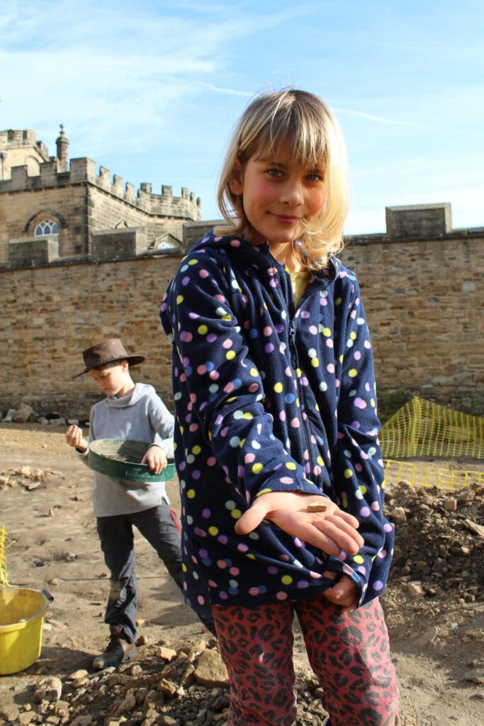 A young person on an archaeological dig site with castle walls in the background holding a small piece of pottery in their hand.