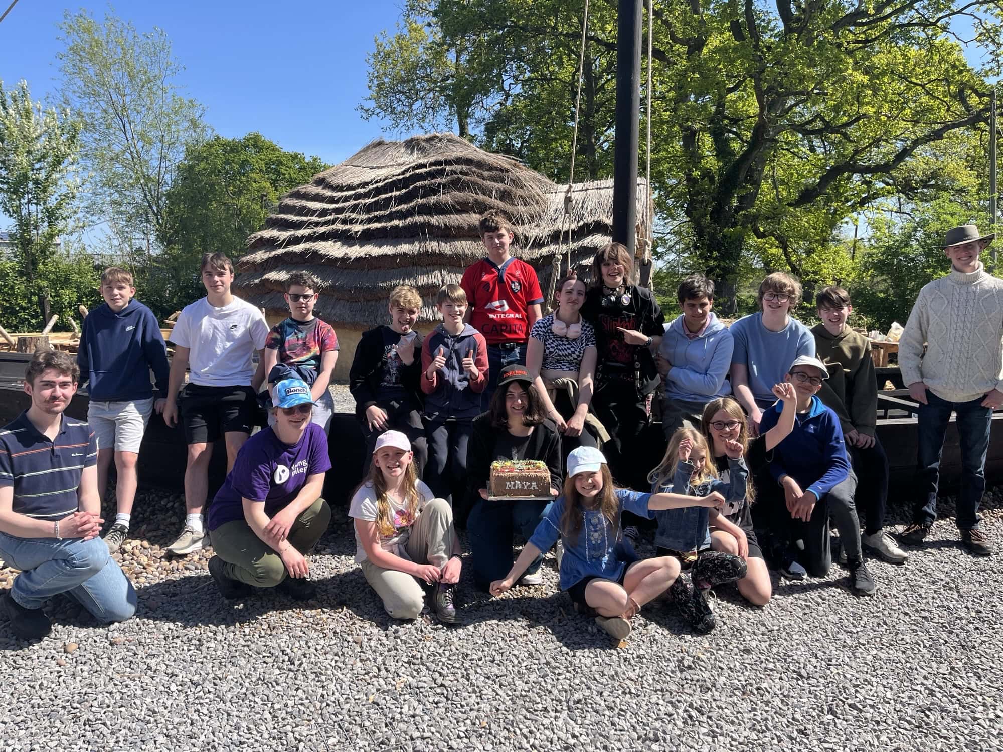 A group of children and adults gathered in front of a reconstructed Anglo-Saxon house on a sunny day.