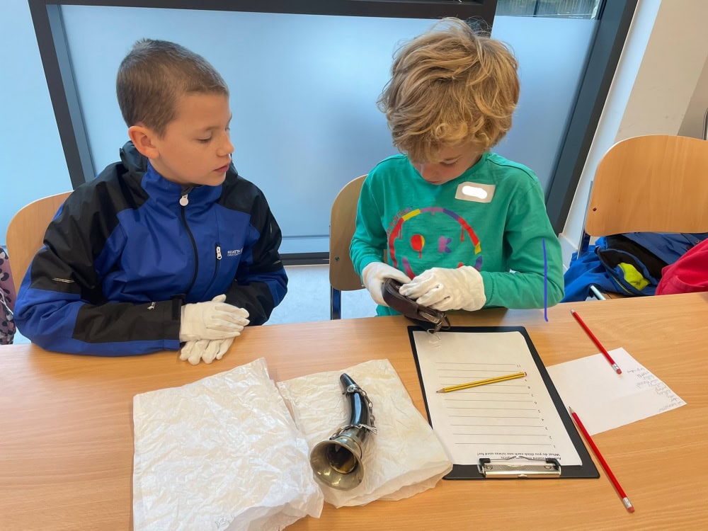 Two young people wearing white gloves sat at a table examining a historic powder horn.