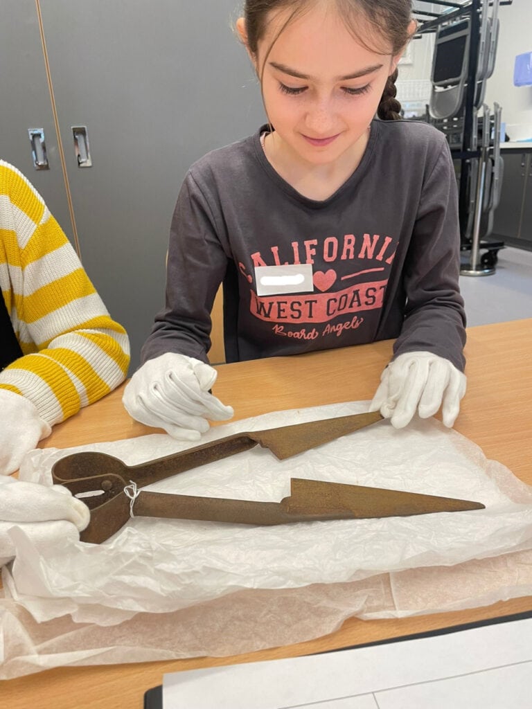 A young person wearing white gloves sat at a table examining a historic pair of shears.