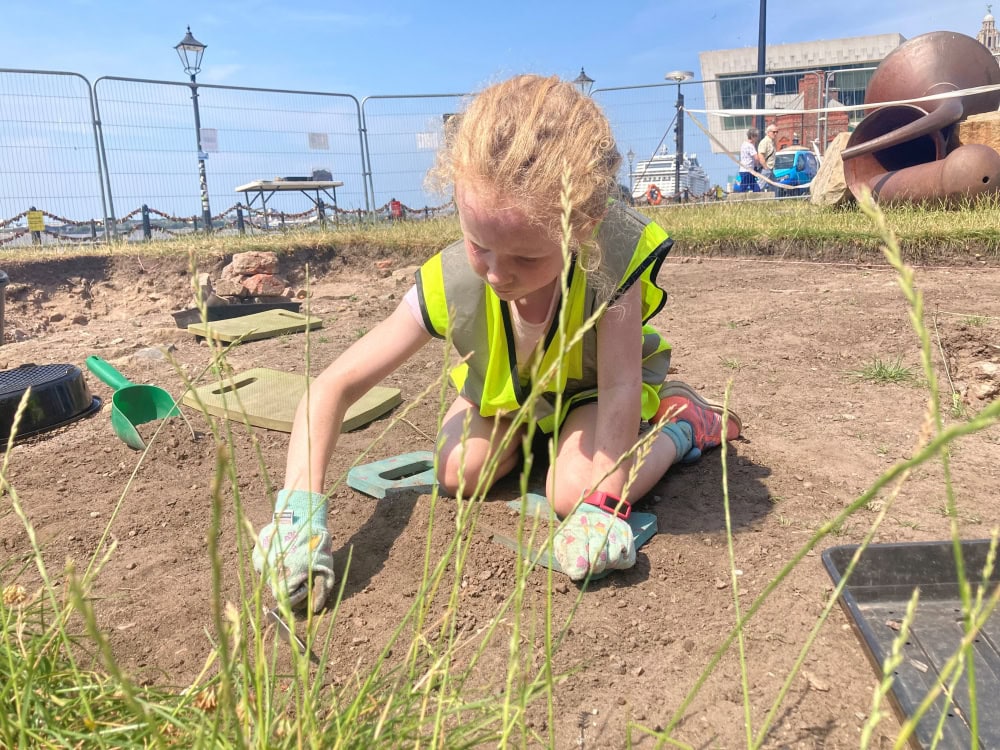 A young person wearing a high-vis vest and gloves kneels in an archaeological trench on a dig and scrapes the soil with a trowel.
