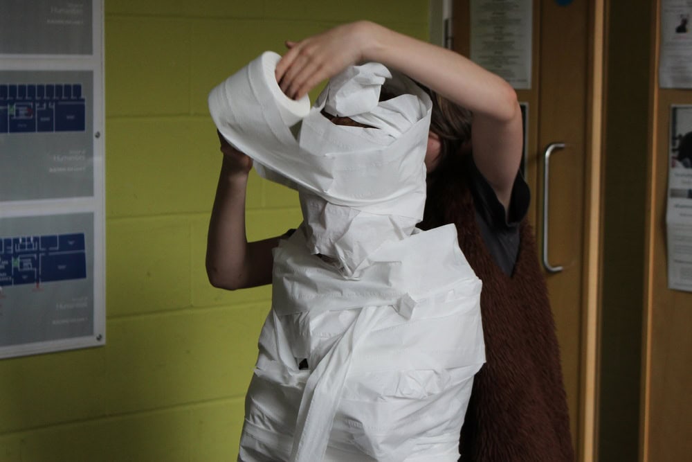 An adult wraps a child entirely with toilet paper so that they resemble an ancient Egyptian mummy.