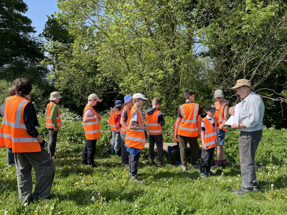 A group of 12 children stand in a group in an outside wooded area whilst an adult talks to them and points to a paper map he is holding.