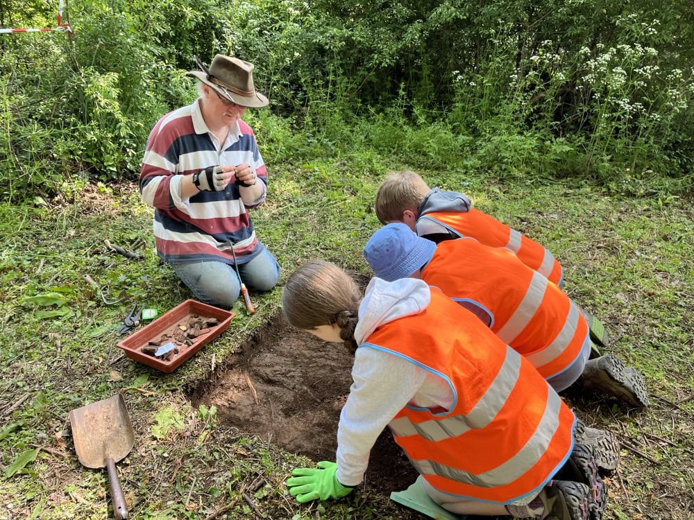 Three children wearing high-vis vests and gloves crouch around the edge of a small shallow square-shaped archaeological trench, which they excavate with trowels. An adult archaeologist kneels at the other end of the trench.