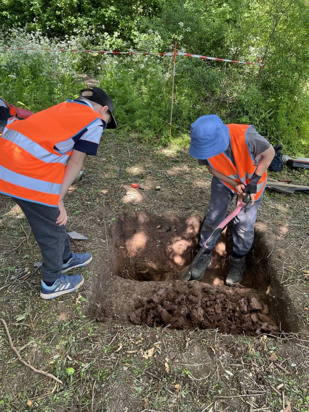 A child wearing high-vis vests and gloves stands in a small square-shaped archaeological trench, which they dig using a spade. Another child leans over the edge to watch.