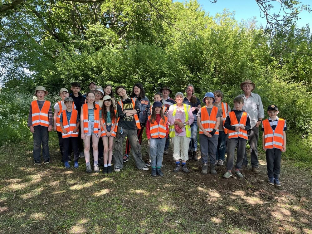 A group of 20 children and 4 adults stand in a group in an outside wooded area.