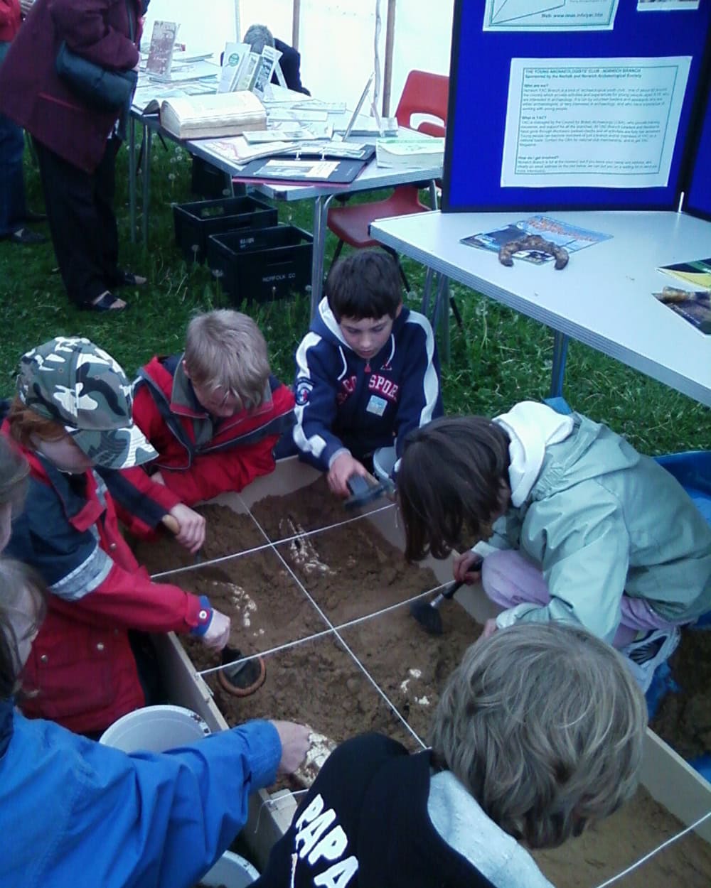 A group of children lean over the edge of a large sandpit to excavate it with brushes and metal trowels.