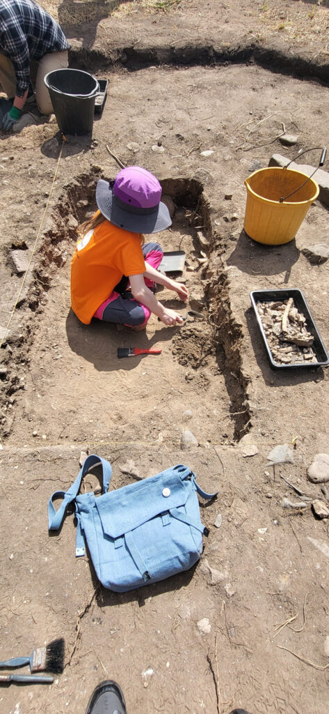 A young person sits in a shallow rectangular trench on an archaeological dig.