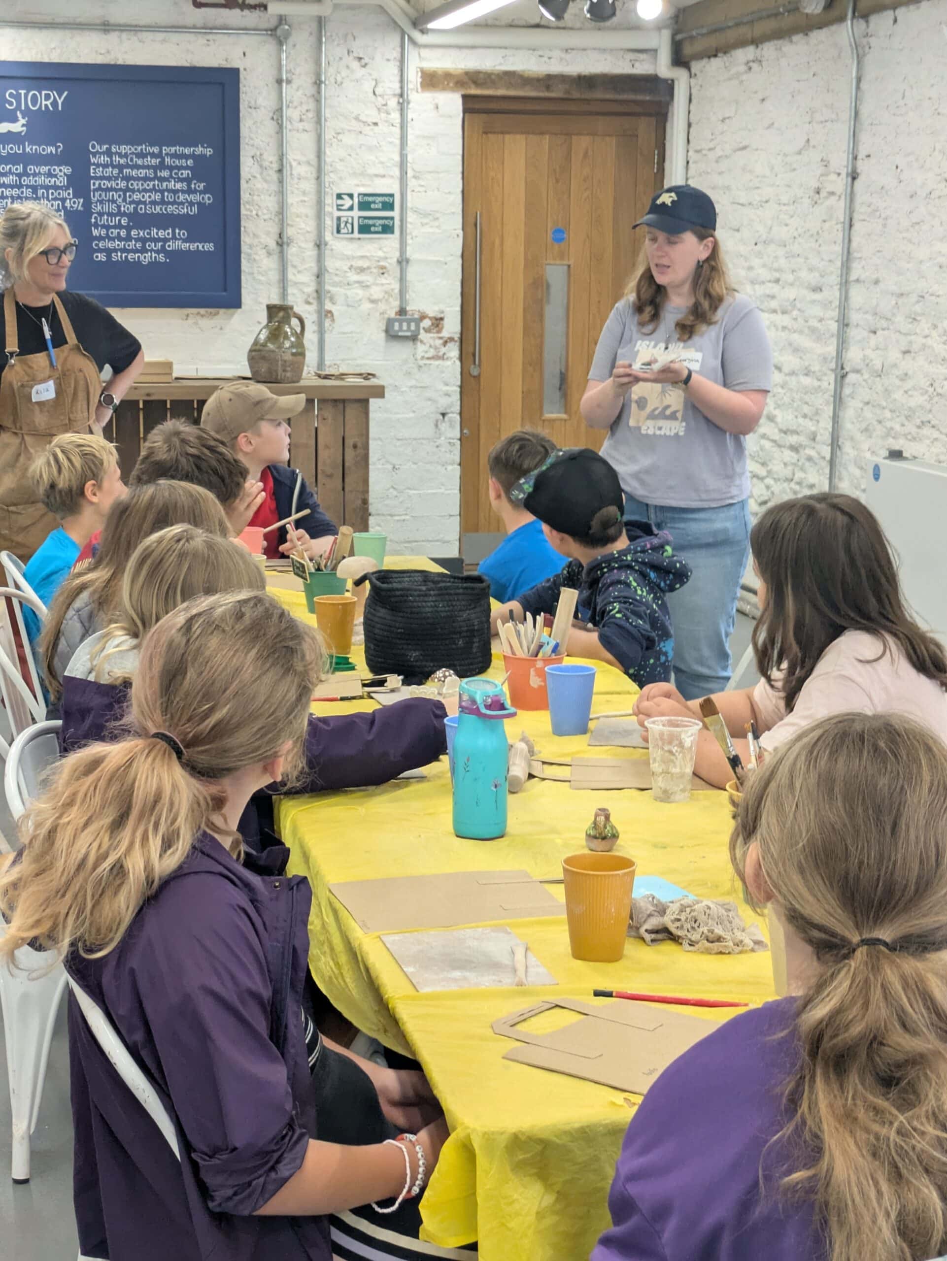 A group of around 10 children are sat around a table with a yellow tablecloth. They are sat listening to an adult wearing a hat explaining something to them.