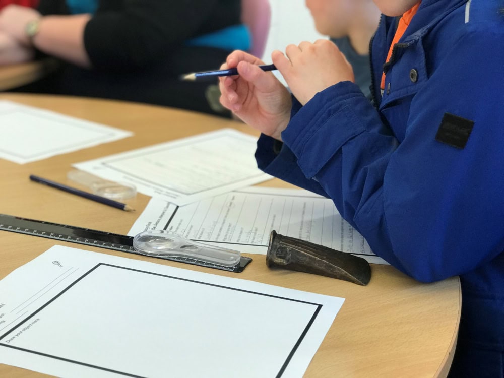 Child sits at a table and completes a fact file worksheet with a pencil. A magnifying glass and a replica bronze axe head are on the table too.