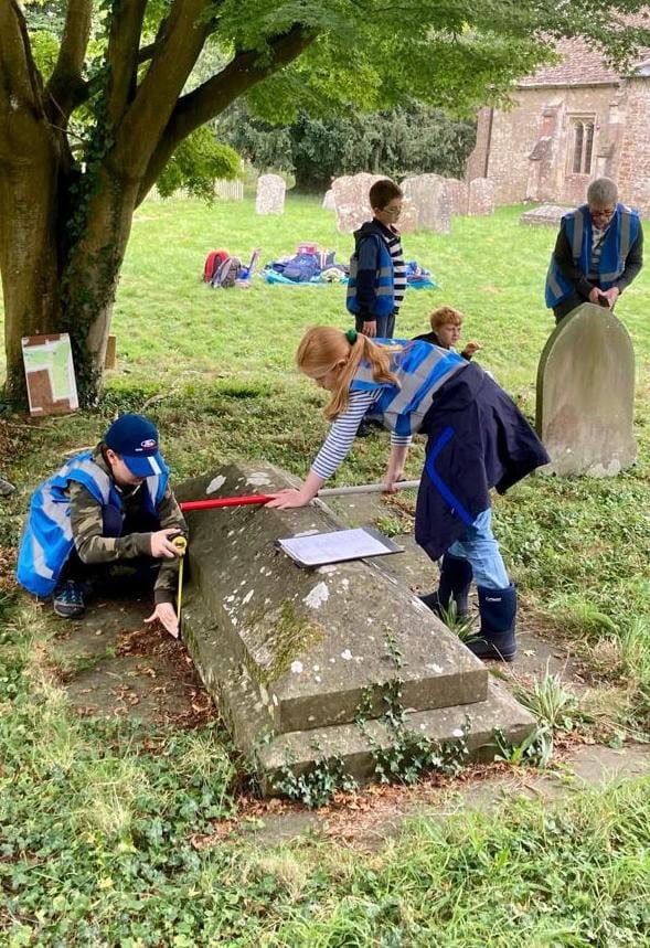 In a church cemetery two young people wearing blue high-vis vests hold a red and white pole over an old tomb stone to measure it.