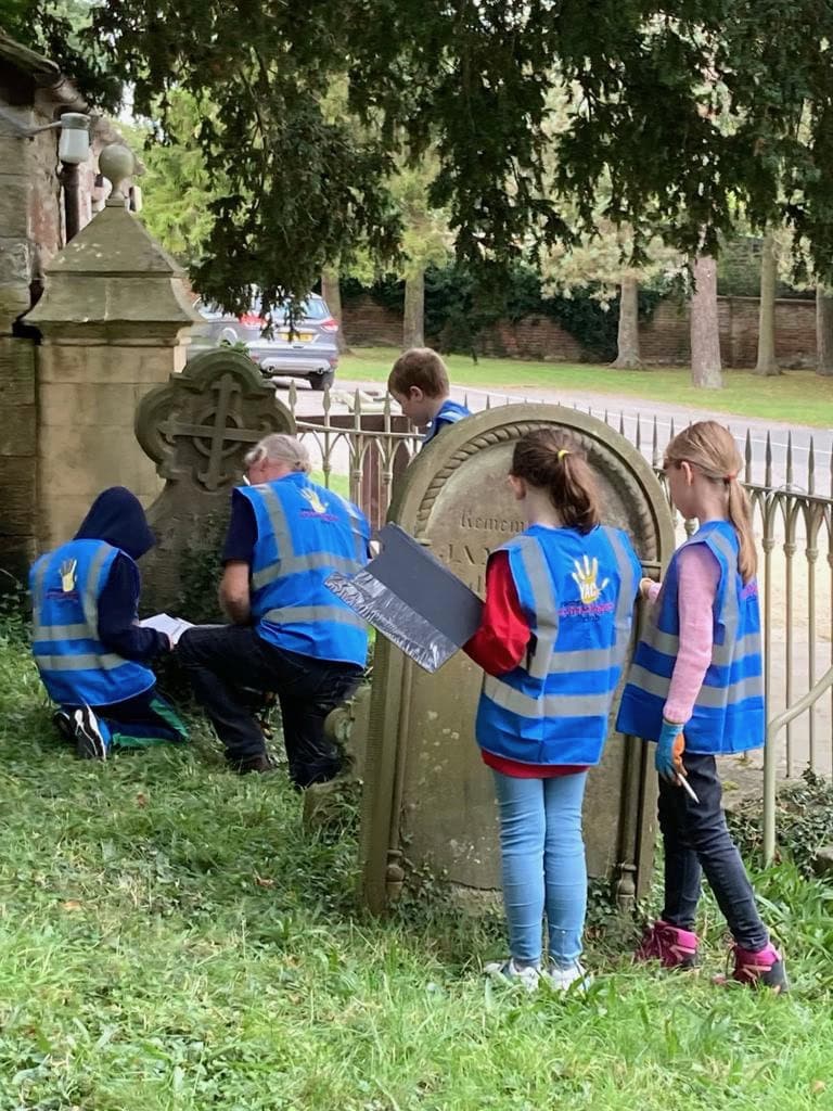 In a church cemetery four young people and one adult wearing blue high-vis vests and holding clipboards stand or crouch in front of old grave stones.