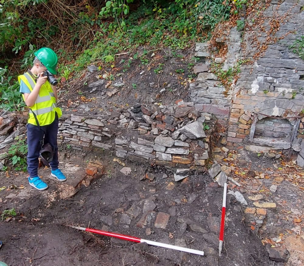 A child wearing a hard hat and high-vis vests photographs an archaeological trench with two red and white ranging rods. In the background is a crumbling stone wall.