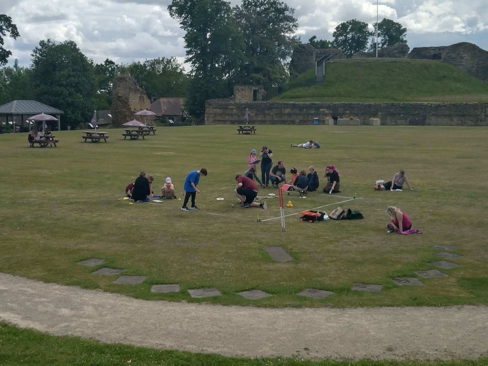 A group of around 20 young people and adults kneeling on a grassy lawn around a marked square.