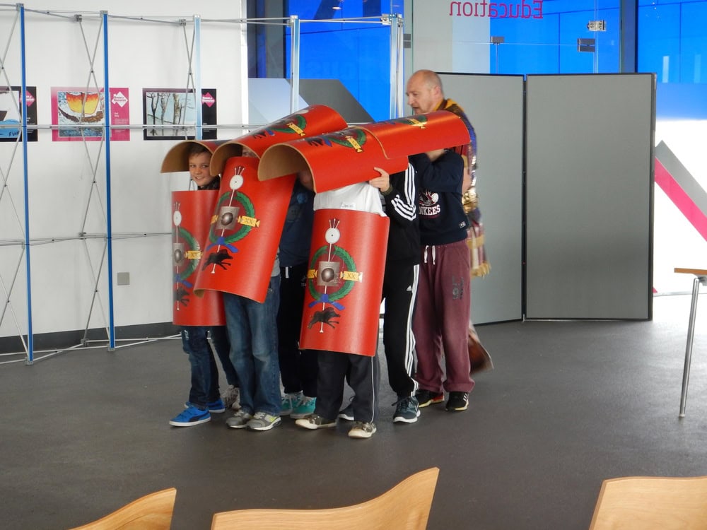 A group of young people carrying replica Roman shields in a formation that covers their heads and bodies.