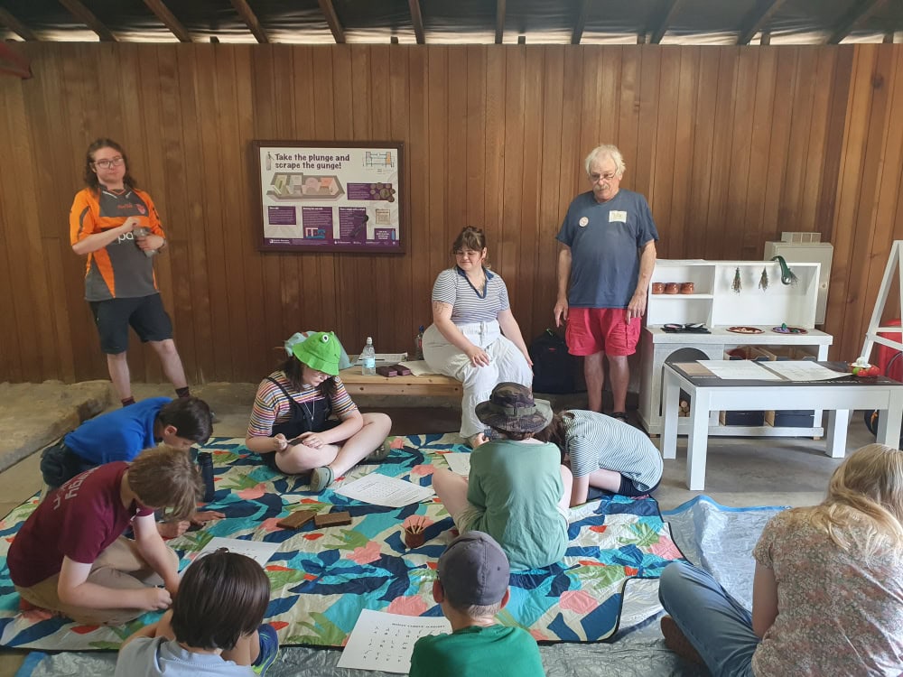 A group of young people sat on a blanket on the floor of a wood-paneled room completing worksheets.