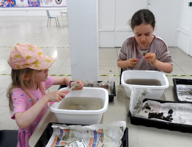 Two children sat at a table washing small archaeological objects in washing-up bowls.