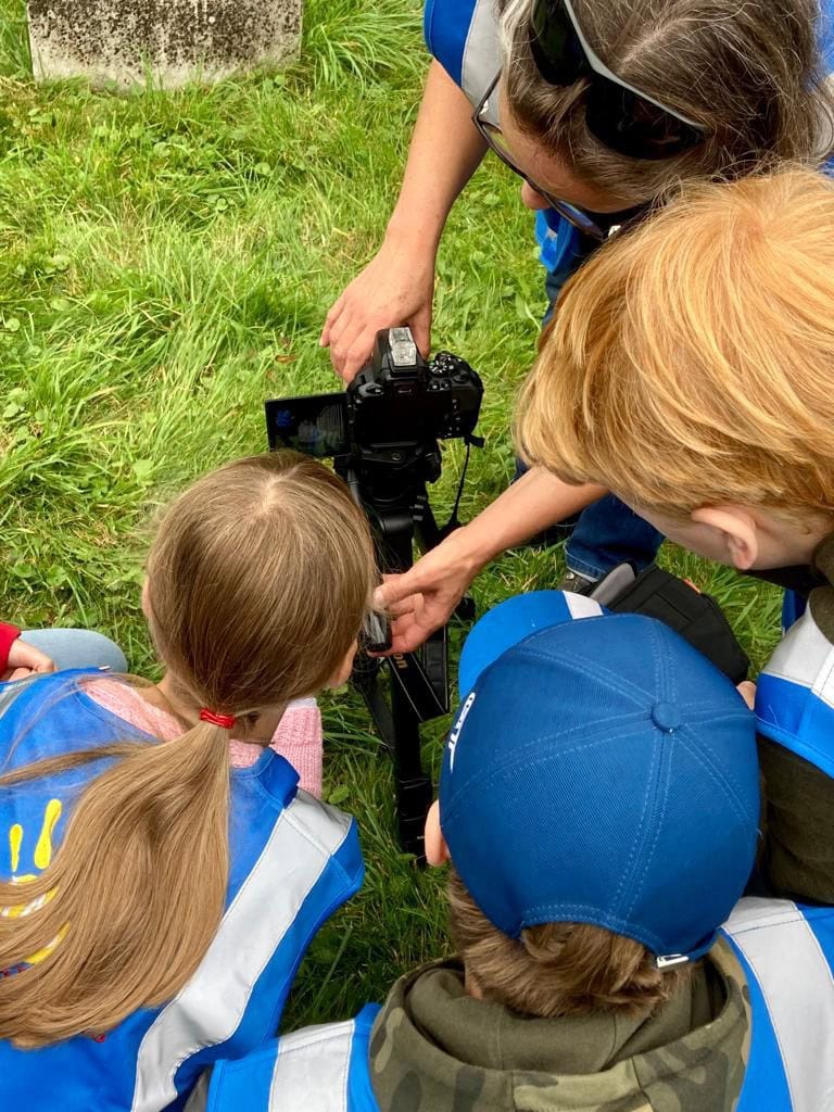 Four young people and one adult wearing blue high-vis vests crouch in front of a professional camera on a tripod on the grass.