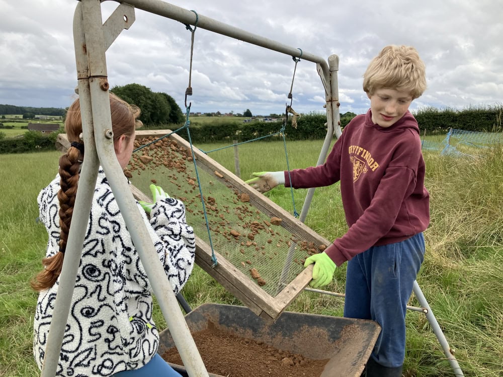 Two young people hold a large flat rectangular sieve of a small archaeological finds over a wheel barrow of soil in a field.