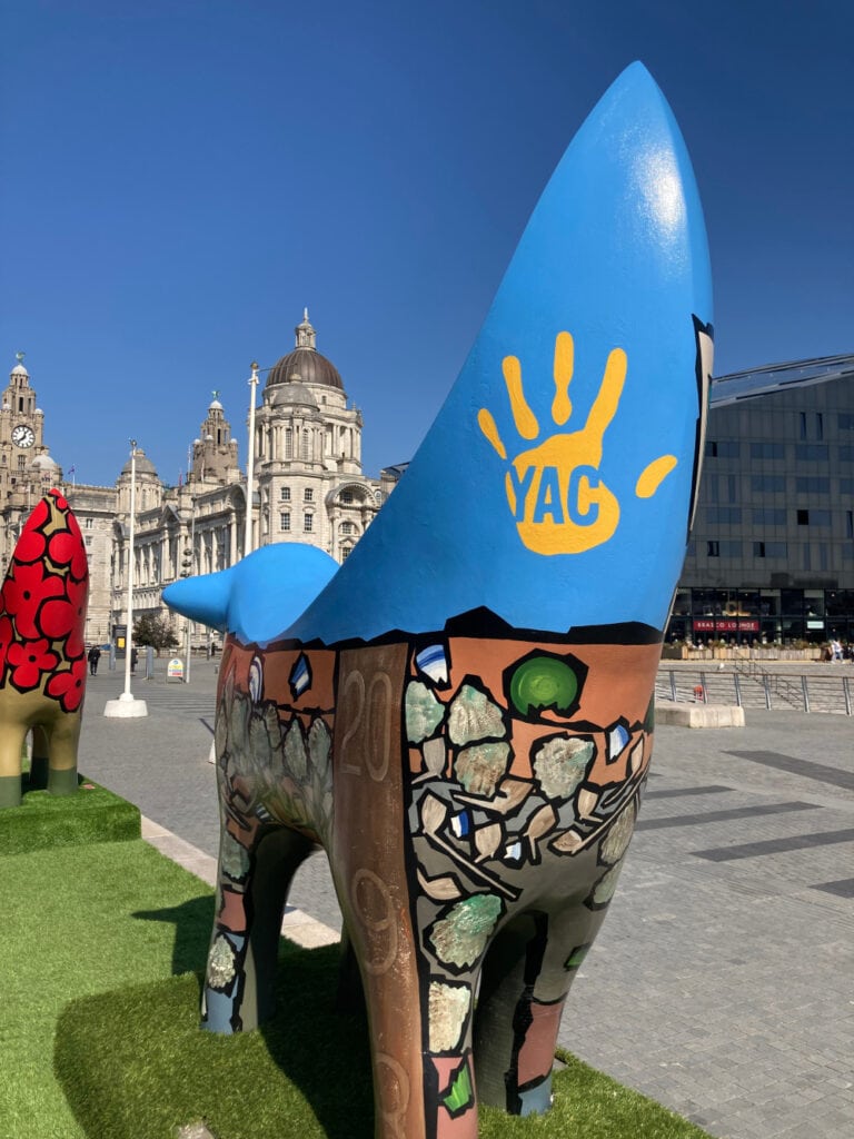An abstract banana-shaped animal stature decorated with a handprint and the YAC logo and other archaeological illustrations such as pottery pieces and shells. The statue is in a city centre.
