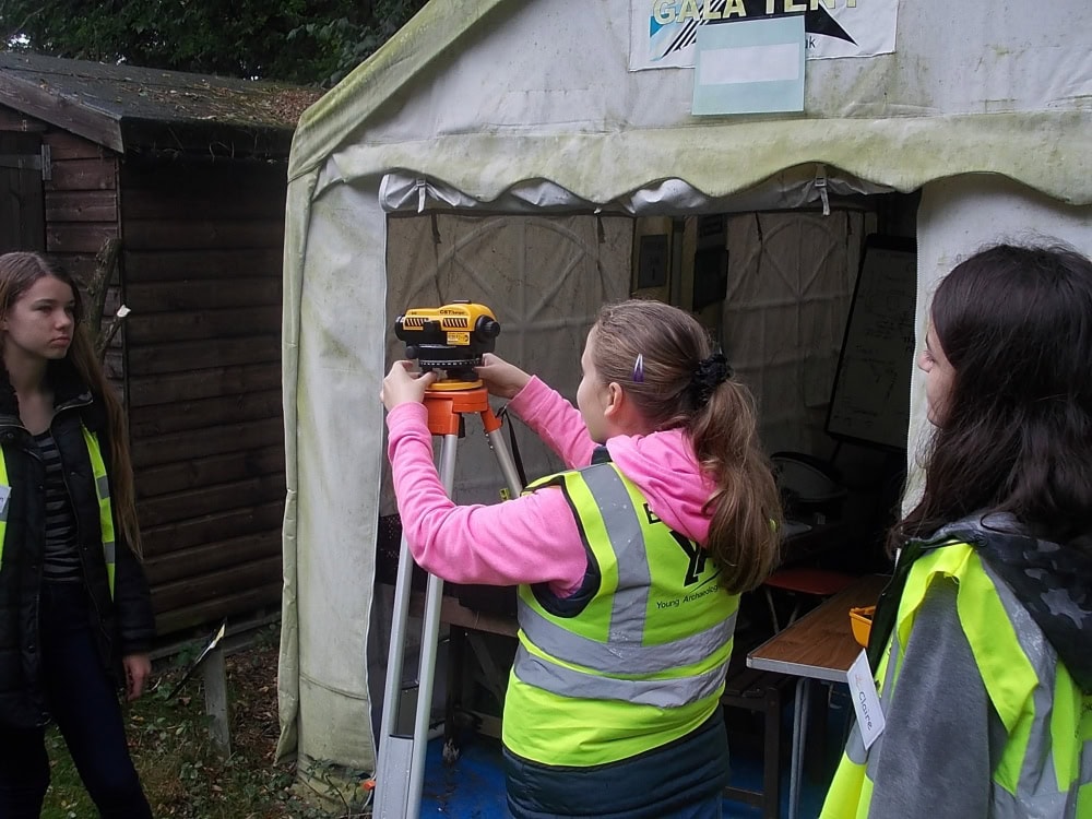 Three young people wearing high-vis vests stand around a dumpy level or piece of surveying equipment on a tripod outside a gazebo.