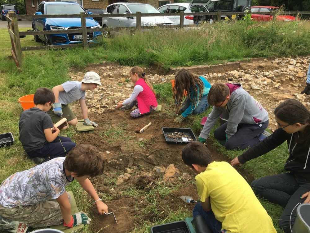 8 young people kneeling around the edge of a shallow rectangular trench which they are excavating with metal trowels.