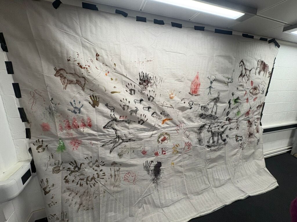 A large sheet pinned to a wall covered in hand prints and drawings in imitation of prehistoric cave art.