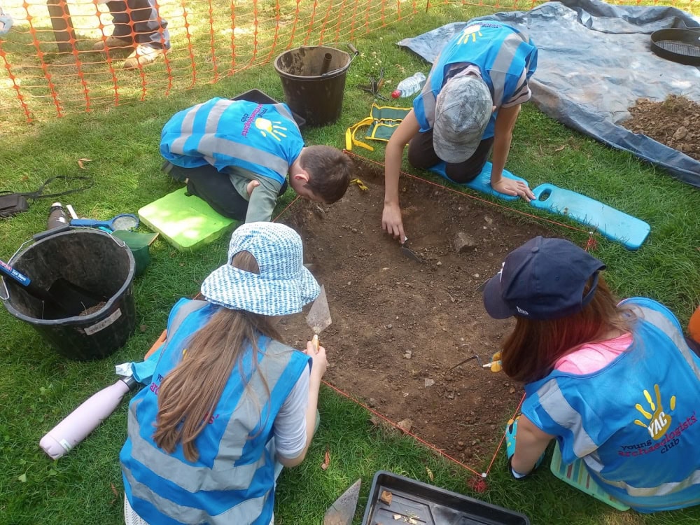 Five young people and one adult wearing blue high-vis vests excavate a small square archaeological trench using trowels.