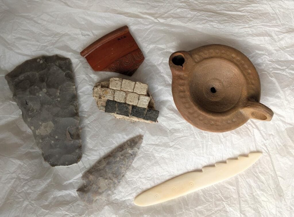 Five artefacts on a piece of white tissue paper: a piece of red Roman pottery, a small clay oil lamp, two flint tools and a piece of craved bone.
