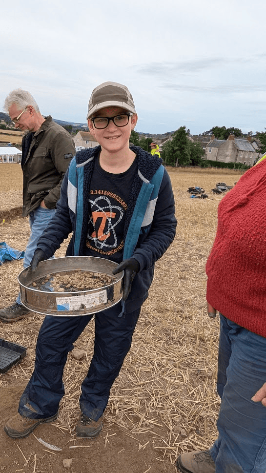 A young person standing in a field on an archaeological dig holding a round sieve.