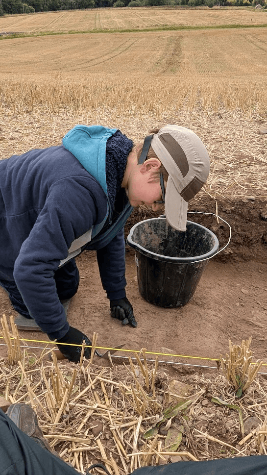 A young person kneeling on the ground in a field scraping the soil with a metal trowel.