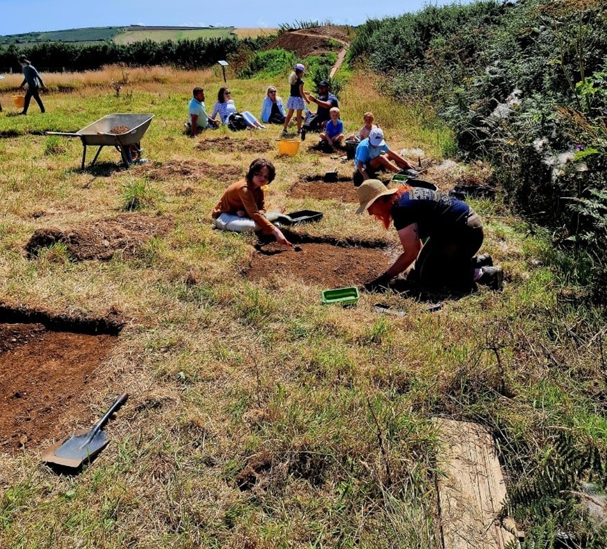 A group of young people on an archaeological dig in the countryside, kneeling between small excavation trenches and wheelbarrows