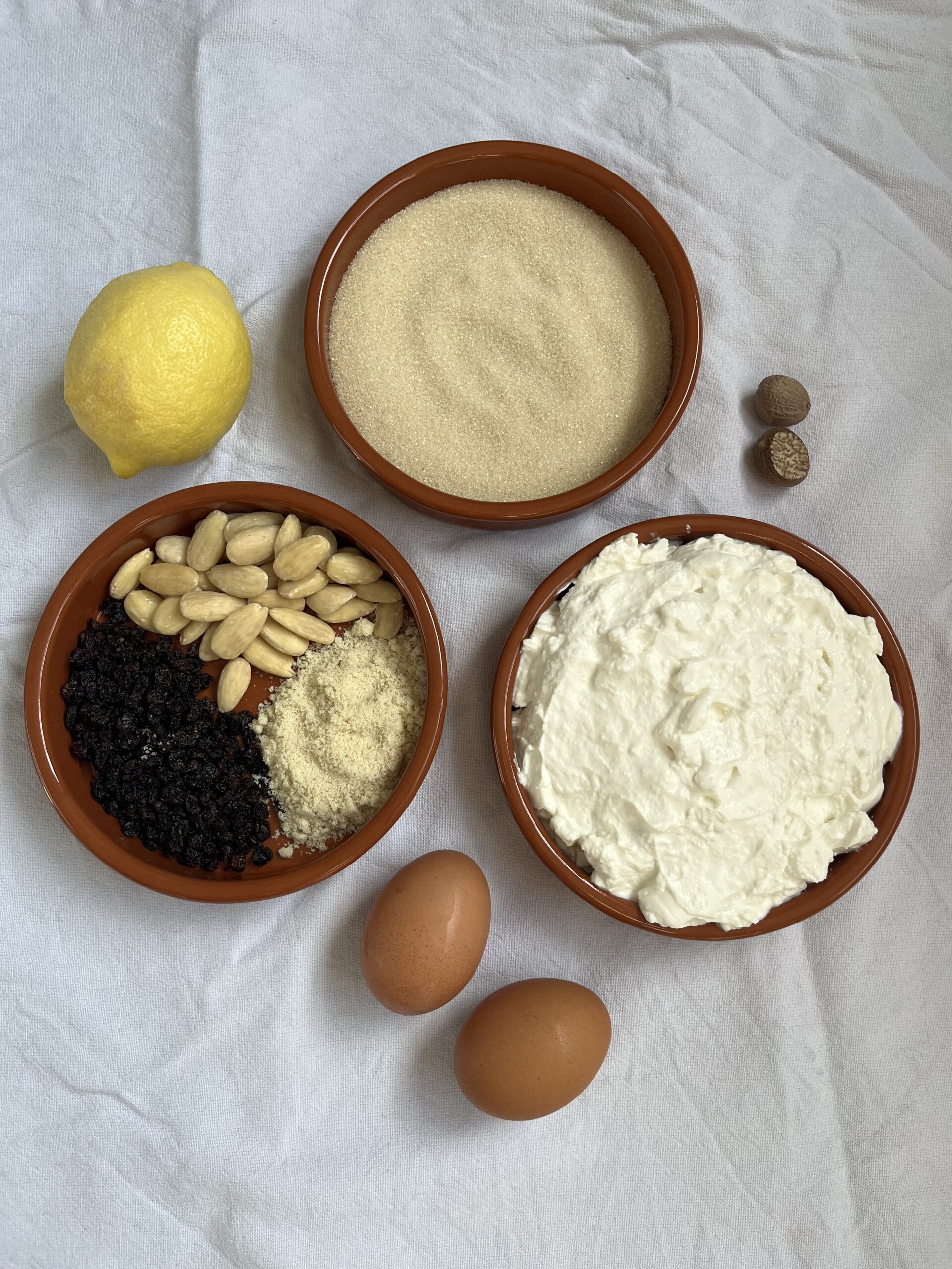 Three small bowls containing sugar, ricotta and a mix of dried currants and almonds, alongside 2 eggs, a lemon and nutmeg.