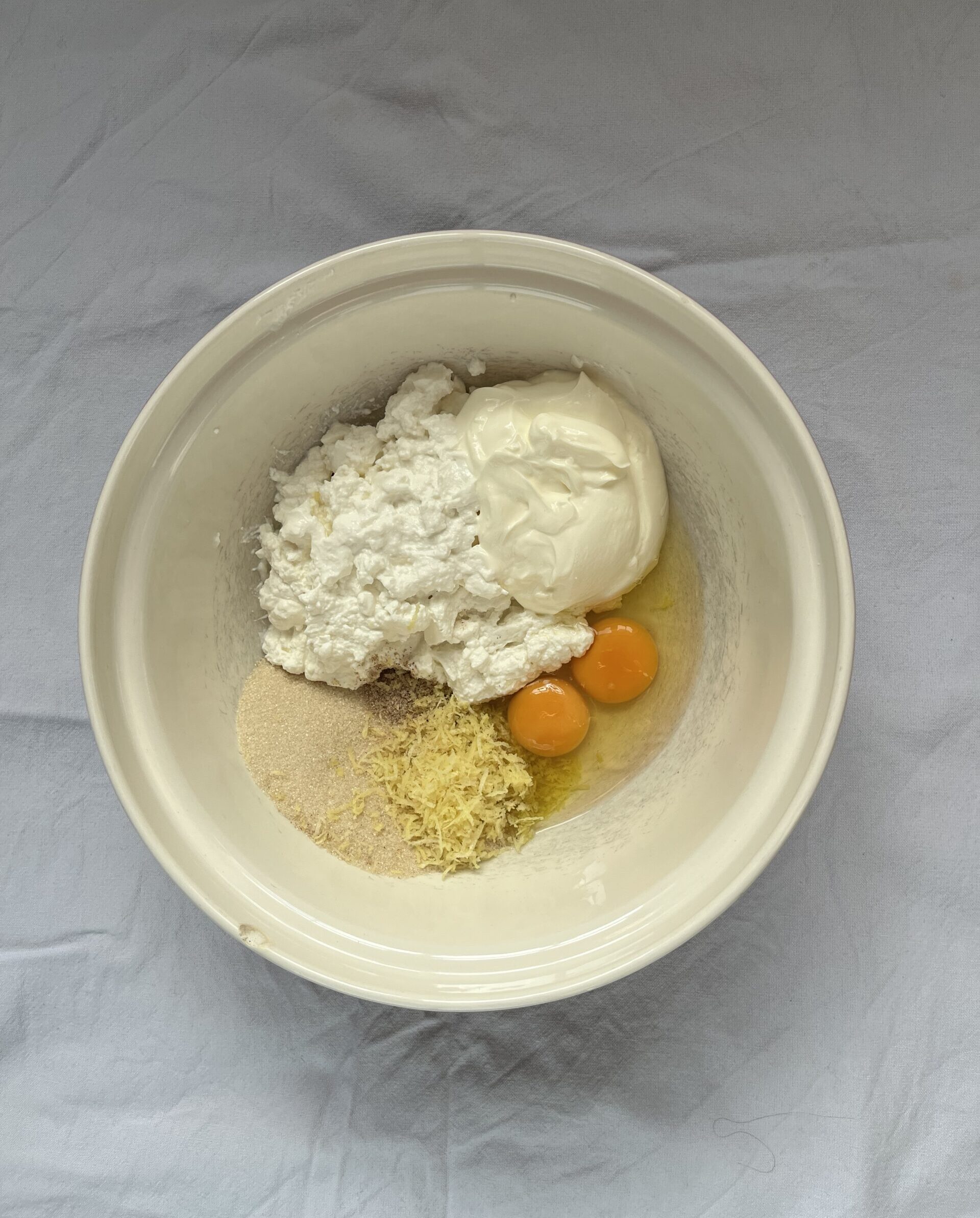 A ceramic mixing bowl containing two raw eggs, sour cream, ricotta, lemon zes and sugar.