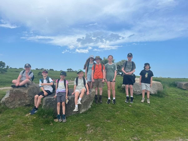Nine young people wearing hats and backpacks stand around large rocks in the middle of a grassy field on a sunny day.