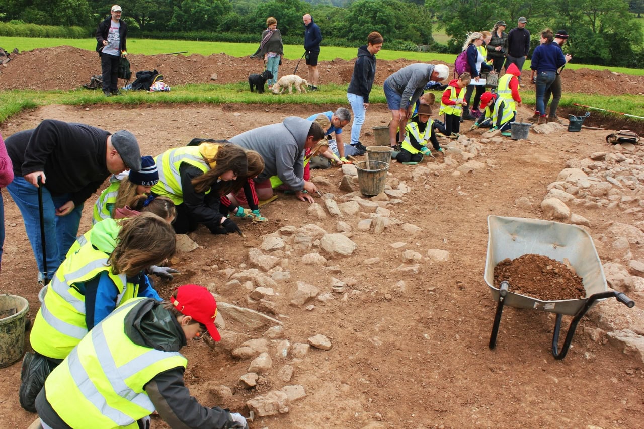 A group of adults and young people digging on an archaeological dig wearing high viz yellow jackets