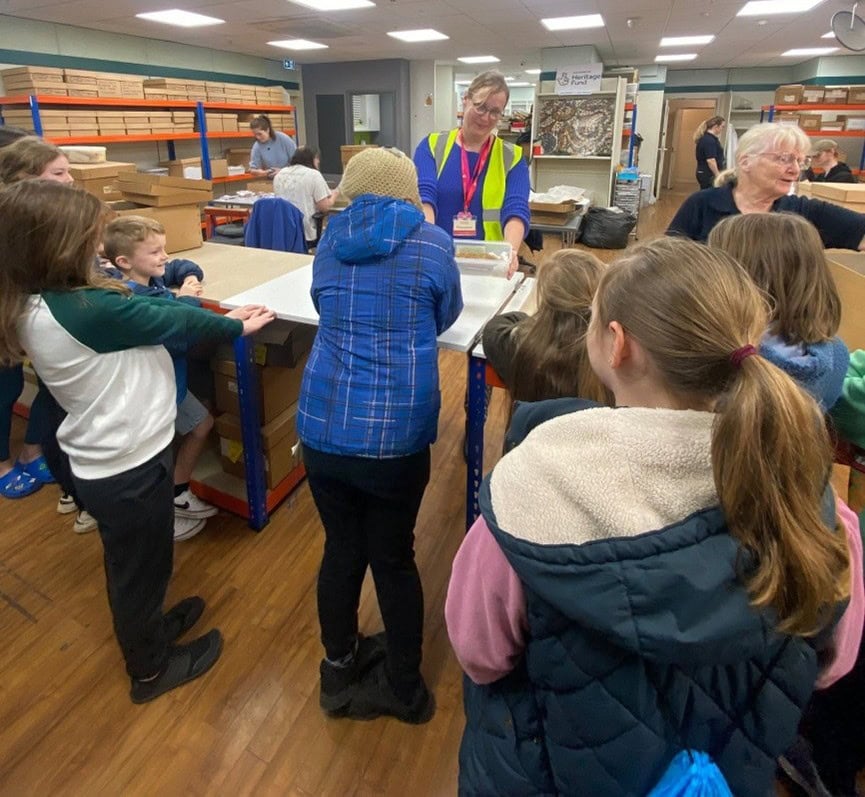 A group of young people gathered around a table in a modern museum archive. An adult standing behind the table shows them a box of objects.