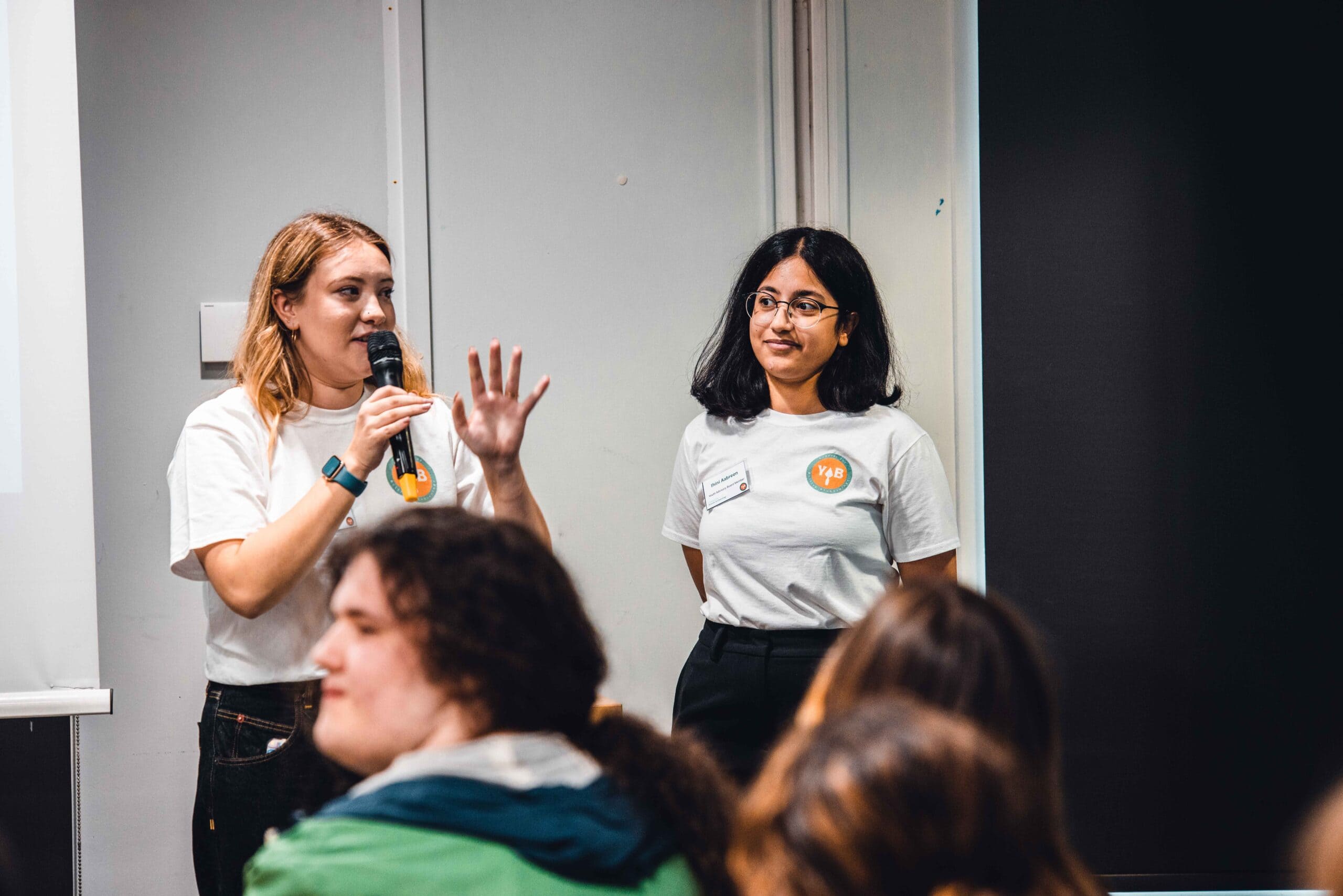 Two people stand at the front of a room, the one of the left is holding a microphone and speaking, the one on the right is listening to what the other person is saying