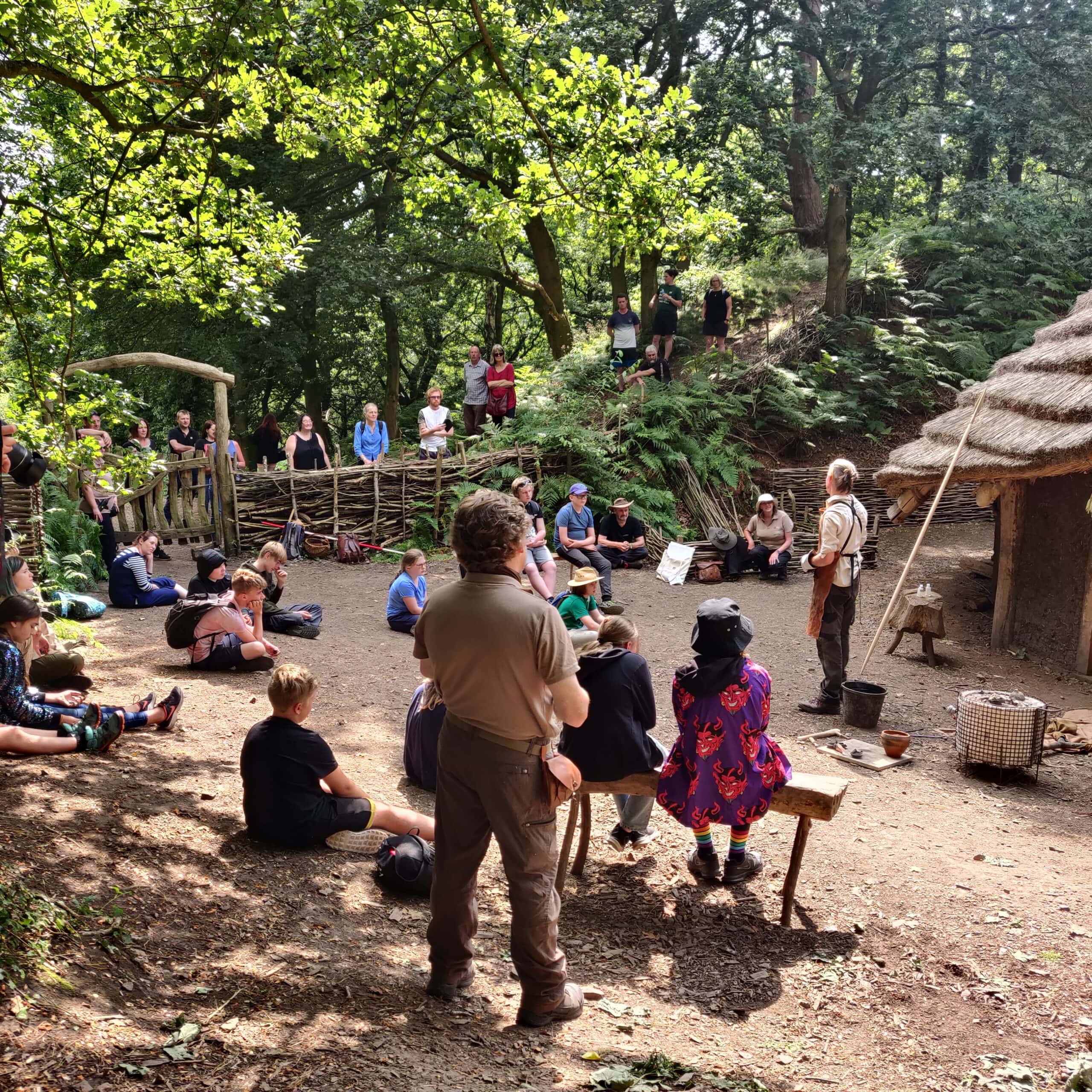 Crowd of people stood in wooded area in front of Bronze Age roundhouse