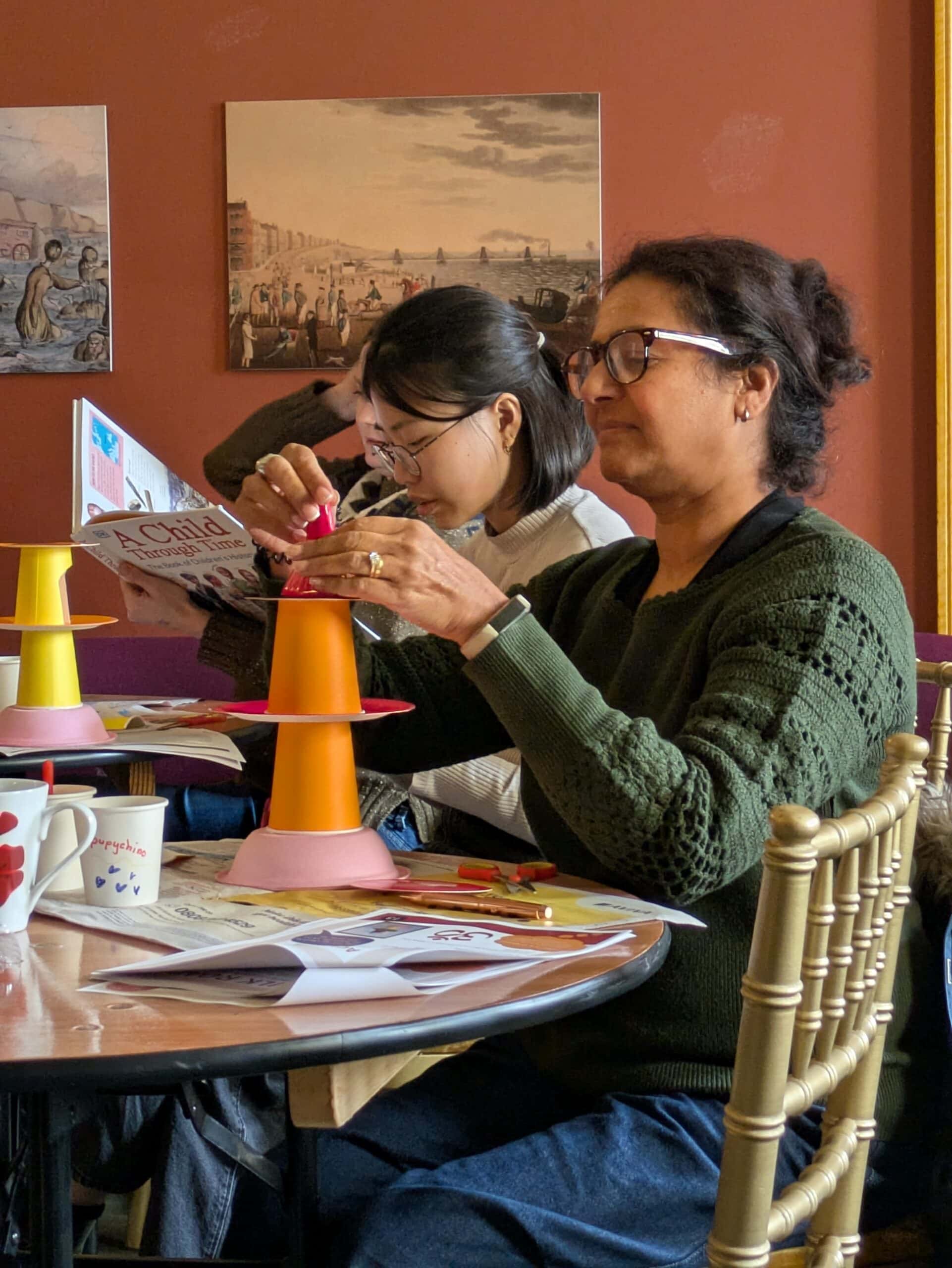 A person wearing a green cardigan sits attempting to build an object. Behind them, there is someone else wearing a white shirt undertaking a different task.
