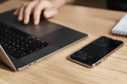 A laptop and phone on a desk, with a hand using the laptop mouse pad
