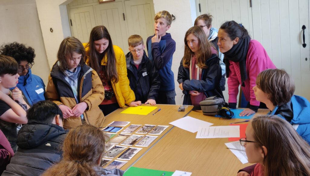 Group of young people stood listening to an adult around a large wooden table with images on it