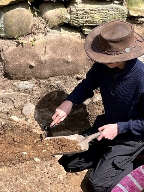 A young archaeologist wearing a brown fedora excavating on an archaeological dig, using a trowel and shovel.