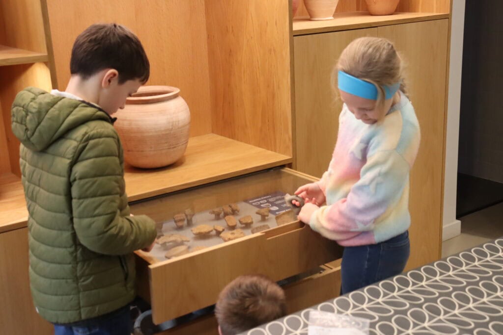 Two young people in a wood-paneled museum exhibition room look at a display case of pottery fragments.